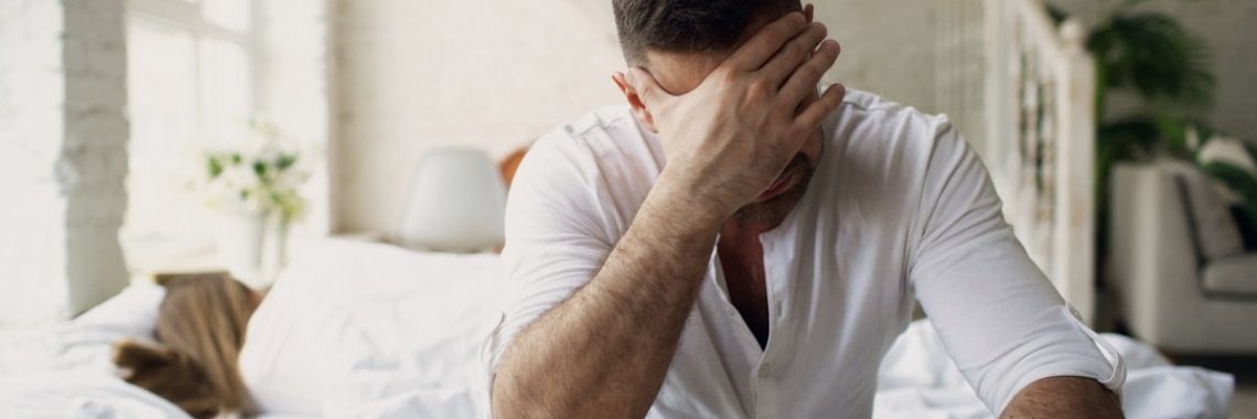 Upset young man sitting in bed have problems with sex while his girlfriend sleep in bedroom in the morning